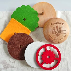 May include: A wooden toy burger set featuring a round bun top engraved with Mama's KITCHEN homemade with love and kitchen utensils, along with felt layers including a green lettuce leaf, orange cheese, brown patty, white onion, and a red tomato slice.