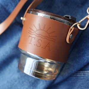 May include: A clear glass tumbler with a brown leather sleeve and a matching strap. The leather sleeve features an engraved sun and wave design with the letters "T.S.A." The strap has gold-colored metal hardware. The tumbler is on a blue denim background.