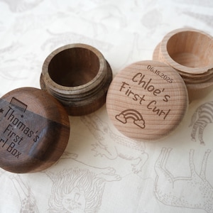 Könnte beinhalten: Vier kleine Holzkästchen, zwei offen und zwei geschlossen, mit graviertem Text. Auf einer Schachtel steht "Thomas's First Curl Box" mit einer Kammgrafik. Eine andere trägt "Chloe's First Curl" mit einem Regenbogen und Datum.