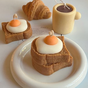 May include: A set of three breakfast-themed candles, including a croissant, a mug of coffee, and a stack of toast with a fried egg on top. The candles are made of a light beige wax and have a realistic appearance.