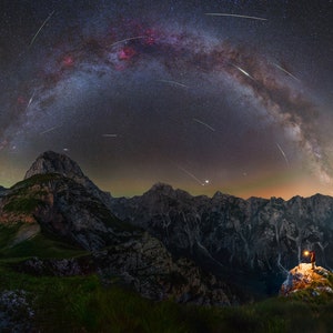 Peut inclure: Paysage nocturne panoramique avec une Voie lactée vibrante au-dessus de montagnes escarpées. Des traînées lumineuses de météores traversent le ciel sombre. Une personne se tient au sommet d'un pic, éclairée par une lumière, ajoutant une échelle.