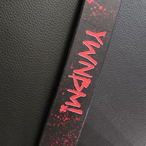 Könnte beinhalten: Ein schwarzes Armband mit einem roten Farbspritzermuster und dem Text YWNBMI in roter Schrift. Das Band ist mit einem schwarzen Druckknopf oben und einem silberfarbenen Metalldruckknopf unten versehen, was einen funktionalen Verschluss bildet.