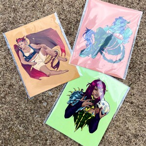May include: Three rectangular art prints in clear protective sleeves. One print features a winged figure with a tail on a tan background. Another shows a blue-skinned figure on a pink background. The third depicts a figure holding a skull on a lime green background.