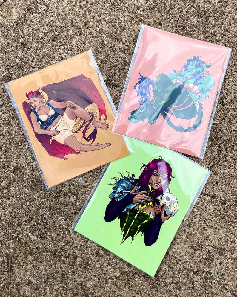 May include: Three rectangular art prints in clear protective sleeves. One print features a winged figure with a tail on a tan background. Another shows a blue-skinned figure on a pink background. The third depicts a figure holding a skull on a lime green background.