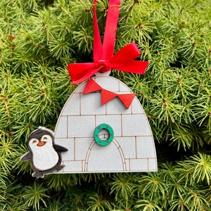 May include: A white wooden ornament shaped like an igloo with a penguin standing outside. The igloo has a green wreath on the door and red pennants hanging from the top. The ornament has a red ribbon attached for hanging.