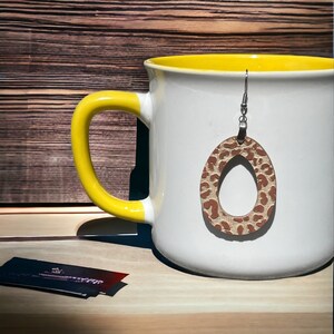 Puede incluir: Una taza de cerámica blanca con asa amarilla y un pendiente de gota con estampado de leopardo marrón y beige colgando del borde.