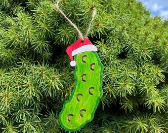 May include: A green pickle Christmas ornament with a red Santa hat. The pickle is made of wood and has a string for hanging.