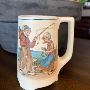 May include: A white ceramic mug with a blue rim and handle. The mug features a hand-painted scene of two children fishing by a lake. The boy is wearing a red shirt and blue pants, and the girl is wearing a purple shirt and blue skirt. The mug is approximately 4 inches tall.