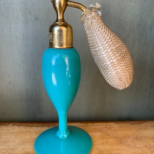 May include: A teal blue glass perfume bottle with a gold top and a fabric spray nozzle. The bottle has a round base and a slender neck.