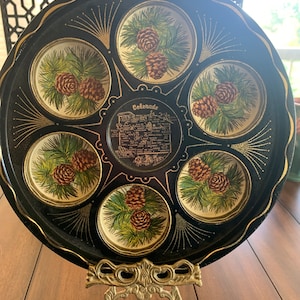 May include: A black metal tray with a gold rim and six circular compartments. Each compartment features a painted image of pine cones and branches. The center of the tray has a gold-colored label with the word "Colorado" and a list of Colorado attractions. The tray is displayed on a gold metal stand.