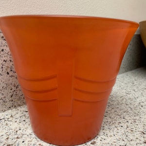 May include: A bright orange ceramic planter with a simple design. The planter has a wide mouth and a rounded bottom.