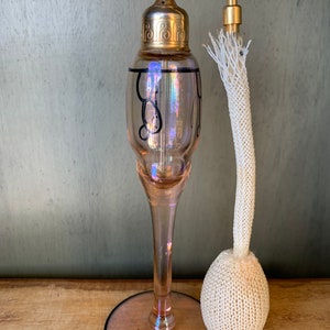 May include: A vintage glass perfume bottle with a gold-toned atomizer and a white crocheted puff. The bottle is pink and has black swirl designs.