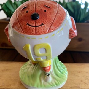 May include: A ceramic figurine of a basketball player wearing a white jersey with the number 19 on the front. The player has a smiling face and is wearing red shorts and white sneakers. The figurine is standing on a green base.