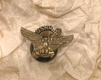 Puede incluir: Un pin de color dorado con un águila y las letras "USNA" en un círculo. El águila sostiene un rollo en su pico.