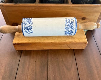 May include: A rolling pin with a central ceramic barrel featuring a white base and blue floral patterns on each end, complemented by light brown wooden handles on both sides. The cylindrical barrel is smooth, while the handles are rounded.