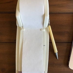 Op de afbeelding: Witte vintage papierdispenser met een rol papier en een pen op een houten oppervlak.