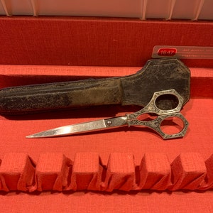 May include: A silver pair of decorative scissors with an ornate design, in a black leather case. The scissors are on a red surface with the text "1847 Rogers Bros. America's First Silverplate" visible.