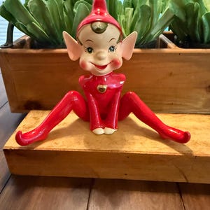 Könnte beinhalten: Vintage Keramik-Elf-Figur mit rotem Outfit und Hut, sitzend mit ausgestreckten Beinen. Der Elf hat ein lächelndes Gesicht, spitze Ohren und ein herzförmiges Detail auf der Brust. Die Figur steht auf einer Holzoberfläche.