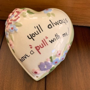 May include: A heart-shaped ceramic trinket box with a floral design and the text "You'll always have a 'pull' with me!"