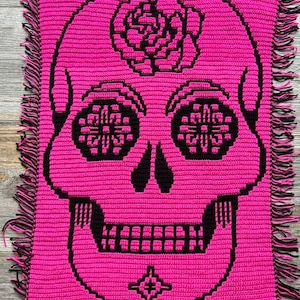 May include: A pink crocheted square with a black sugar skull design. The skull has a flower on its forehead and intricate designs on its eyes.