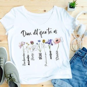 May include: White v-neck t-shirt with the text "Dieu dit que tu es" and floral illustrations. The flowers are in various colors, including pink, purple, and blue. The shirt is paired with jeans and sneakers.