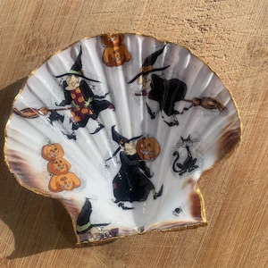 May include: A white scallop shell dish with a gold-painted rim, featuring a repeating Halloween-themed pattern of cartoon witches in black hats, orange jack-o'-lantern pumpkins, and a black cat. The interior surface is smooth and glossy.