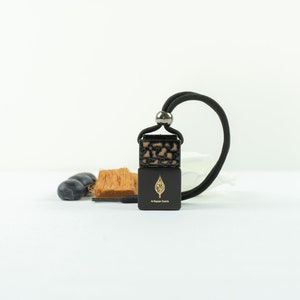 Puede incluir: Un ambientador de coche negro con un diseño de hoja dorada y el texto "The Fragrance Bar" colgando de una cuerda negra.
