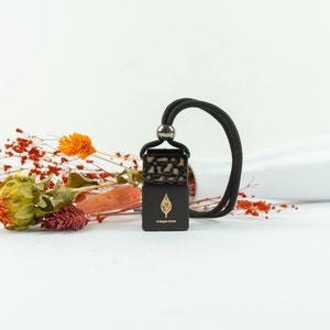 Puede incluir: Ambientador de coche negro con un logotipo de hoja y el texto "in upper earth". El ambientador tiene un cordón negro y una parte superior estampada. Flores secas y un jarrón blanco en el fondo.