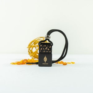May include: A black car air freshener with a gold leaf design and a black cord. The air freshener is sitting on a white surface with small pieces of amber scattered around it.