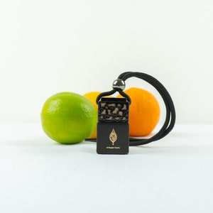 Puede incluir: Un ambientador de coche negro con un diseño de hoja dorada que cuelga de un cordón negro. El ambientador está delante de una lima y una naranja.