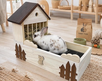May include: A wooden rabbit feeder and litter box combination featuring a house-shaped hay rack with a dark brown roof, light wood walls, decorative dark brown picket fence accents, and a small wooden rabbit cutout on the front panel.
