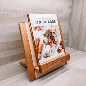 May include: A wooden cookbook stand with a book titled "ON BOARDS" is displayed. The stand is made of light and dark wood, with a slanted design to hold the book upright. The book cover features images of food.