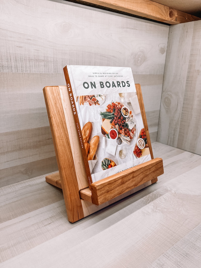 May include: A wooden cookbook stand with a book titled "ON BOARDS" is displayed. The stand is made of light and dark wood, with a slanted design to hold the book upright. The book cover features images of food.