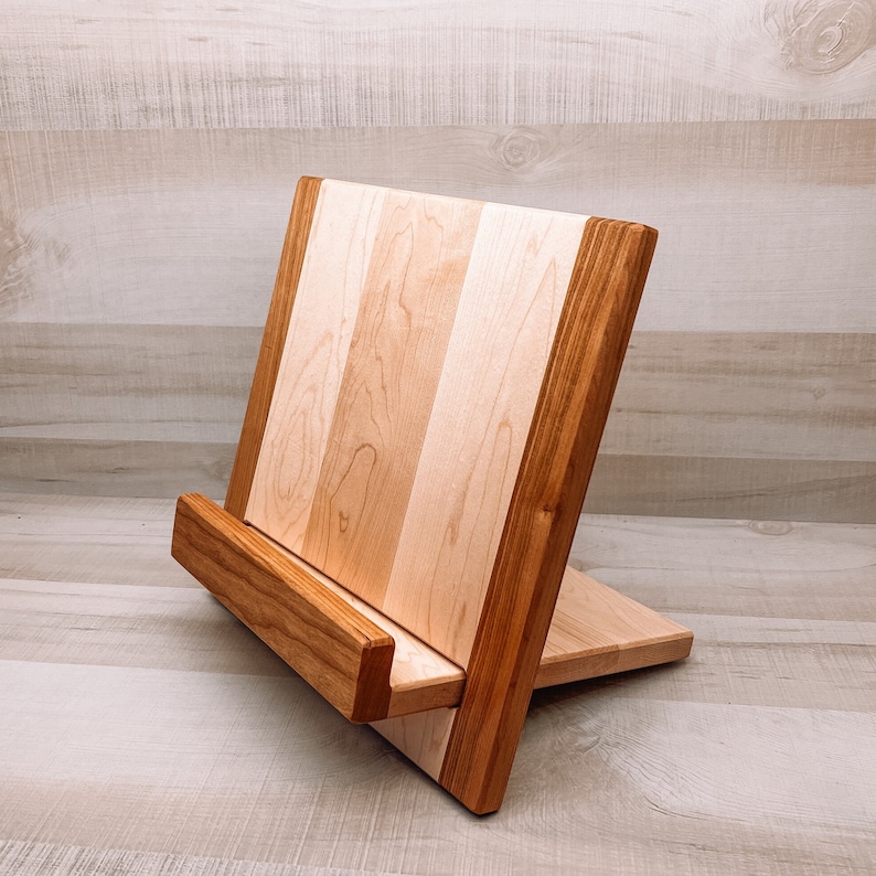 May include: A wooden book stand with a light-colored, striped center and darker brown edges. The stand is angled for easy viewing and has a small ledge to hold books. The wood grain is visible, giving it a natural, rustic look.