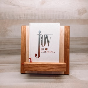 May include: A wooden cookbook stand with a book titled "JOY OF COOKING." The stand is made of light and dark wood, with a small shelf to hold the book. A red ribbon marks a page.