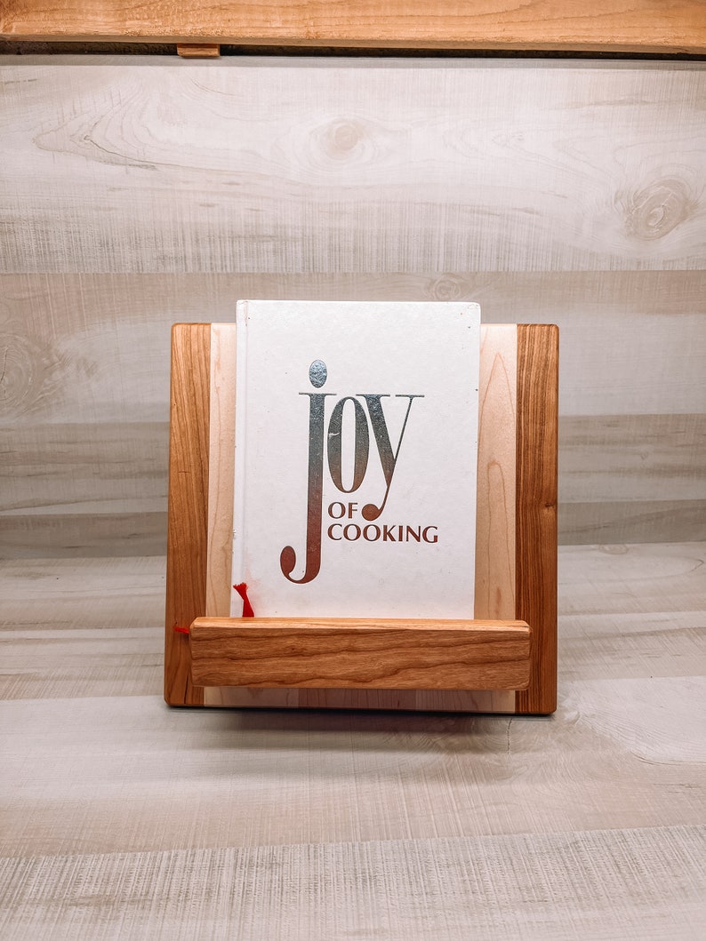 May include: A wooden cookbook stand with a book titled "JOY OF COOKING." The stand is made of light and dark wood, with a small shelf to hold the book. A red ribbon marks a page.