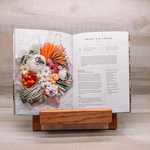 May include: An open cookbook titled "Bright & Blooming" displays a colorful arrangement of vegetables. The book rests on a wooden book stand. The vegetables include carrots, cauliflower, and tomatoes. The book stand is made of light-colored wood.