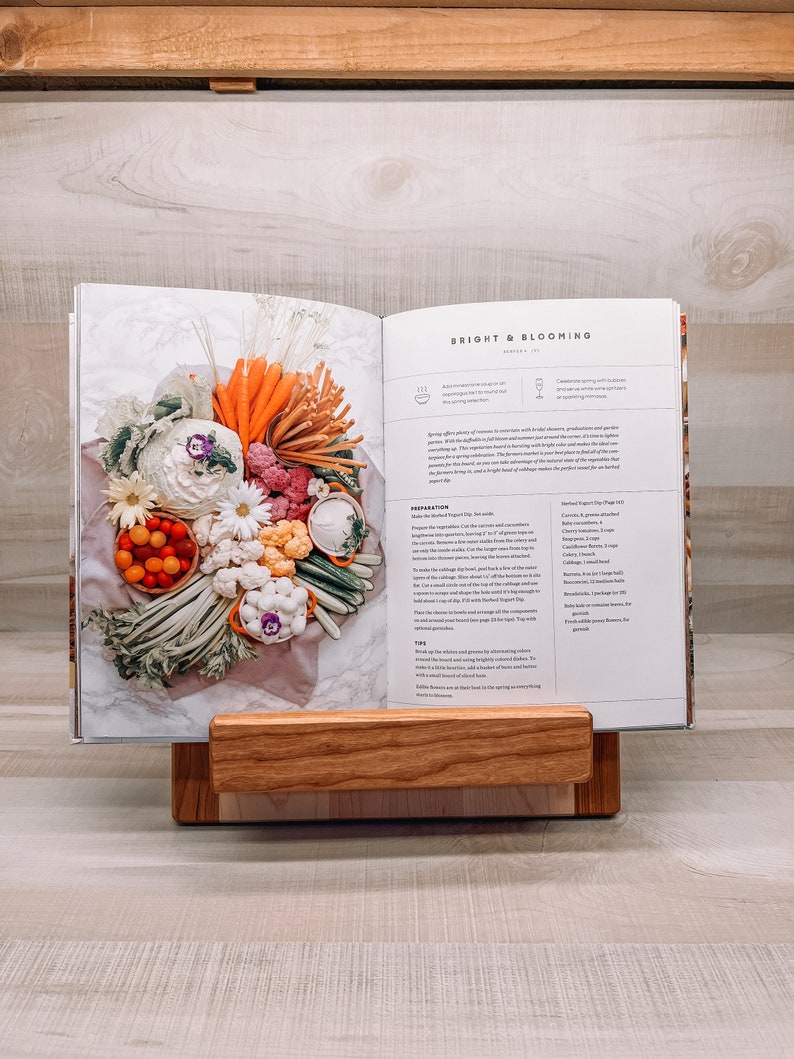 May include: An open cookbook titled "Bright & Blooming" displays a colorful arrangement of vegetables. The book rests on a wooden book stand. The vegetables include carrots, cauliflower, and tomatoes. The book stand is made of light-colored wood.