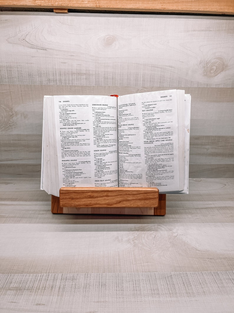 May include: A wooden cookbook stand holds an open cookbook, displaying recipes. The stand is made of light-colored wood and has a horizontal ledge to support the book. The cookbook is open to a page with text.