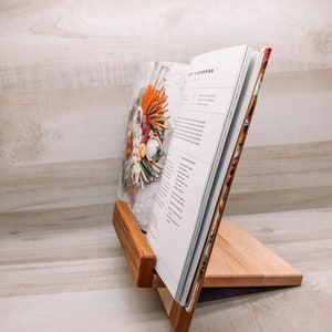 May include: A wooden cookbook stand holds an open book with the title "Light & Blooming." The stand is made of light-colored wood with a darker wood accent. The book's cover features a floral arrangement.