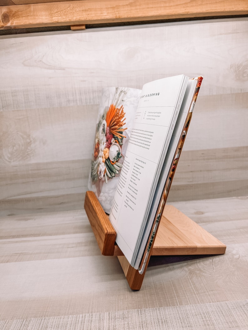 May include: A wooden cookbook stand holds an open book with the title "Light & Blooming." The stand is made of light-colored wood with a darker wood accent. The book's cover features a floral arrangement.