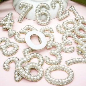 May include: A set of pearl and rhinestone embellishments shaped like numbers. The numbers are white with a gold outline and are arranged on a pink surface.