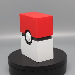 May include: A rectangular storage box featuring a red top half and a white bottom half, separated by a black horizontal band. A circular white button is centered on the black band, mimicking the design of a classic red and white ball container.