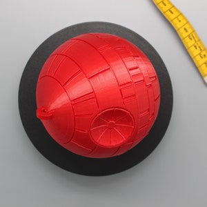 Pode incluir: Modelo esf&eacute;rico vermelho que se assemelha a uma Estrela da Morte, repousando sobre uma base circular preta. O modelo tem detalhes de superf&iacute;cie. Uma fita m&eacute;trica amarela &eacute; parcialmente vis&iacute;vel. Item de colecionador.