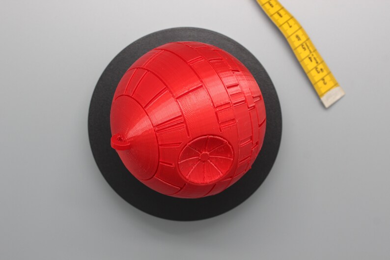 Pode incluir: Modelo esf&eacute;rico vermelho que se assemelha a uma Estrela da Morte, repousando sobre uma base circular preta. O modelo tem detalhes de superf&iacute;cie. Uma fita m&eacute;trica amarela &eacute; parcialmente vis&iacute;vel. Item de colecionador.