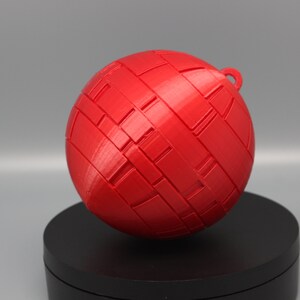 Pode incluir: Uma esfera vermelha vibrante com um padr&atilde;o tecido e uma pequena argola no topo. A bola decorativa &eacute; provavelmente impressa em 3D e assenta numa base preta, perfeita para decora&ccedil;&atilde;o de casa ou como ornamento.