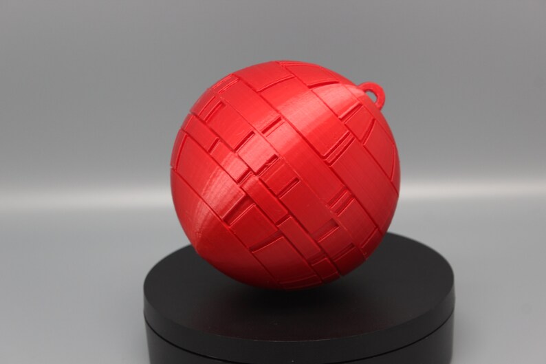 Pode incluir: Uma esfera vermelha vibrante com um padr&atilde;o tecido e uma pequena argola no topo. A bola decorativa &eacute; provavelmente impressa em 3D e assenta numa base preta, perfeita para decora&ccedil;&atilde;o de casa ou como ornamento.