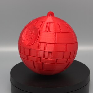 Pode incluir: Um ornamento esf&eacute;rico vermelho, que se assemelha &agrave; Estrela da Morte de um filme de fic&ccedil;&atilde;o cient&iacute;fica. O ornamento tem uma argola para pendurar e &eacute; constru&iacute;do com pain&eacute;is retangulares. Um design circular &eacute; vis&iacute;vel em um lado.