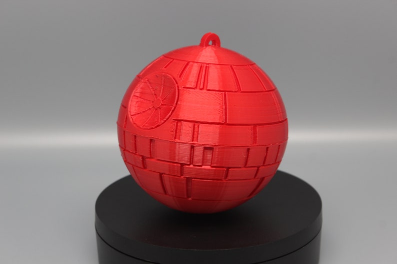 Pode incluir: Um ornamento esf&eacute;rico vermelho, que se assemelha &agrave; Estrela da Morte de um filme de fic&ccedil;&atilde;o cient&iacute;fica. O ornamento tem uma argola para pendurar e &eacute; constru&iacute;do com pain&eacute;is retangulares. Um design circular &eacute; vis&iacute;vel em um lado.