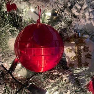 Pode incluir: Um enfeite de Natal vermelho em forma de Estrela da Morte pendurado numa &aacute;rvore de Natal nevada. O enfeite &eacute; uma esfera com detalhes na superf&iacute;cie. Um enfeite branco em forma de presente com detalhes dourados tamb&eacute;m &eacute; vis&iacute;vel.